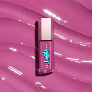 Frasco de gloss ou batom líquido em tom rosa vibrante, com tampa metálica prateada, deitado sobre uma superfície com textura cremosa da mesma cor. O frasco possui uma arte gráfica azul e branca com letras estilizadas.