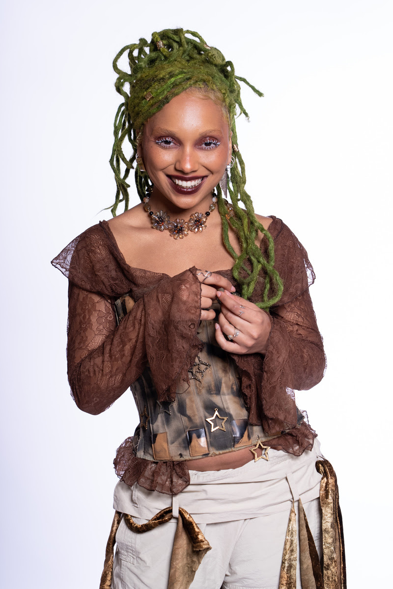 Pessoa negra sorridente com cabelo verde em dreadlocks presos no alto da cabeça. Ela usa uma blusa de renda marrom com mangas longas e largas, um corset estampado com estrelas douradas e calça clara com detalhes dourados. Usa colar com flores e maquiagem expressiva com batom escuro e cílios destacados.