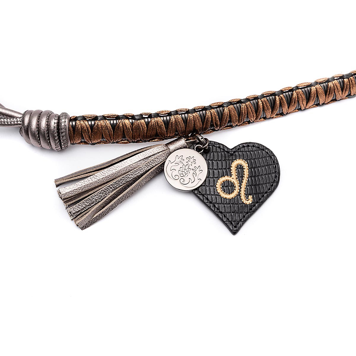 Detalhe de um acessório com pingentes. Ele possui uma tira trançada em tons de cobre e preto, um pingente metálico prateado com desenho floral, uma franja metalizada e um pingente em forma de coração preto com textura, contendo o símbolo do signo de Leão em dourado.