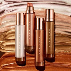 Quatro frascos de iluminador corporal líquidos da marca Fenty Beauty em tons perolado, dourado, bronze e cobre estão dispostos em pé sobre uma superfície texturizada com camadas de produto espalhado nas mesmas cores. As embalagens são elegantes, com tampa metálica rosé gold e o nome "Fenty Beauty" gravado verticalmente nos frascos translúcidos.